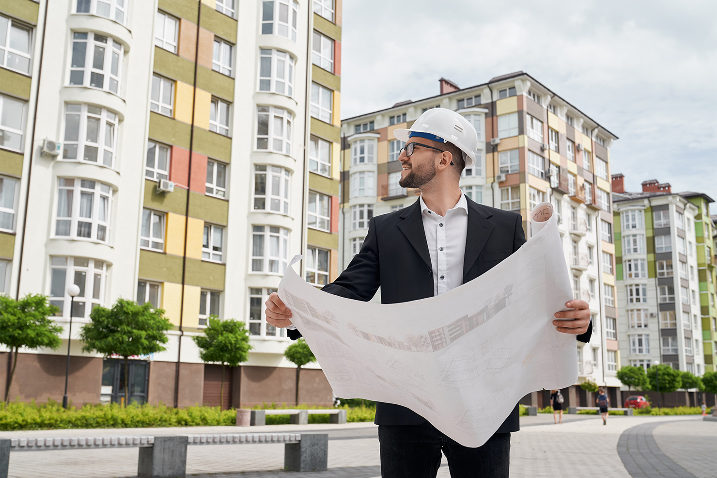 Inspector tehnic purtând cască de protecție și analizând planurile de construcție în fața unui bloc de locuințe, ilustrând serviciile de administrare tehnică oferite de Urban Hive Admin.